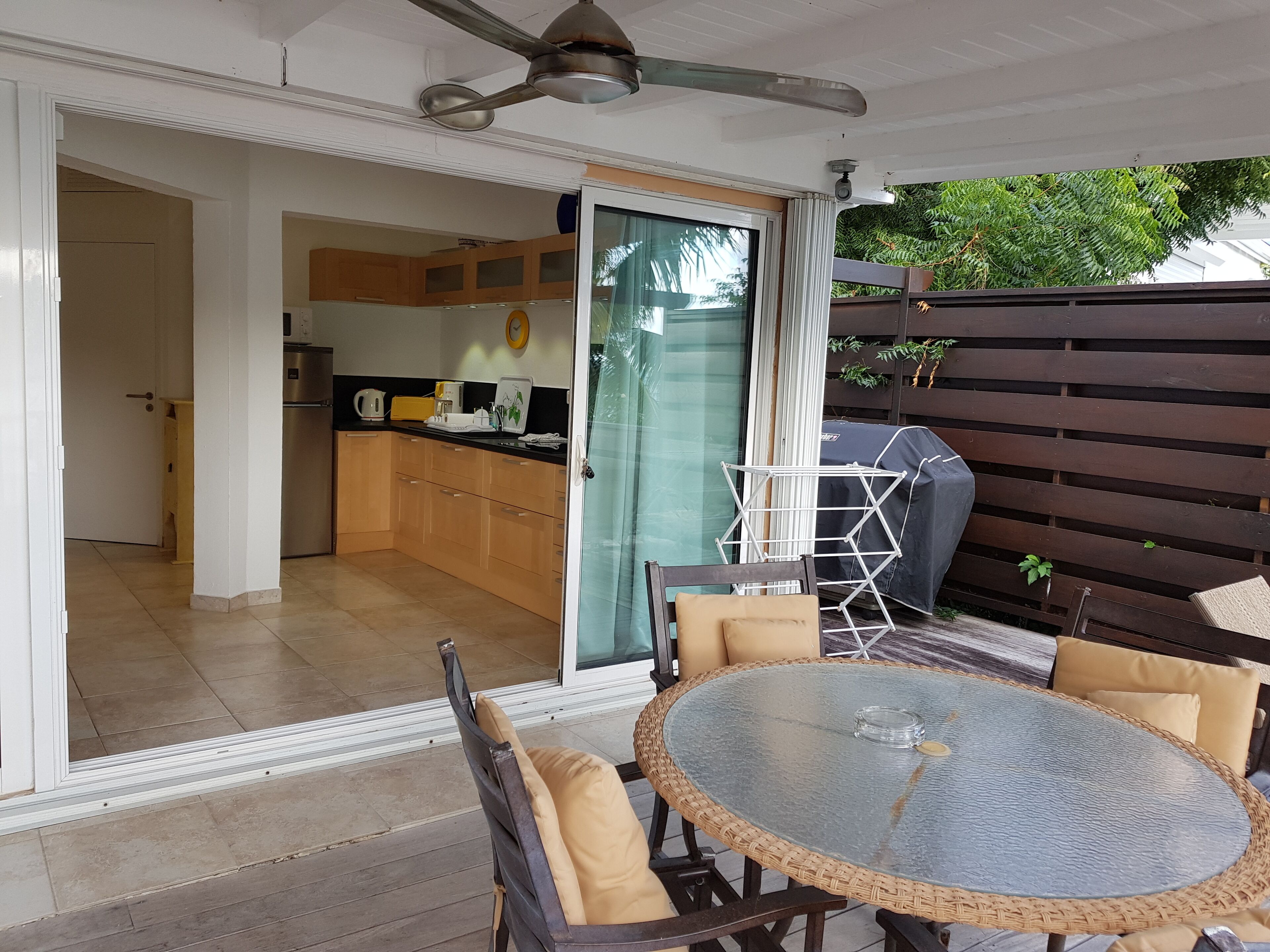 A patio with a round glass table and wicker chairs, a ceiling fan, and a view of the kitchen and garden.