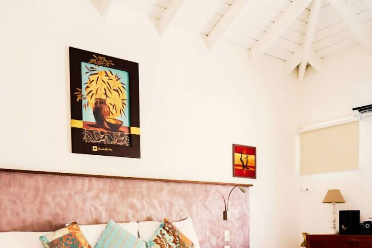 A neatly made bed with a pink bedspread and multiple pillows, a wooden headboard, a wall-mounted lamp, and a framed artwork above the bed.