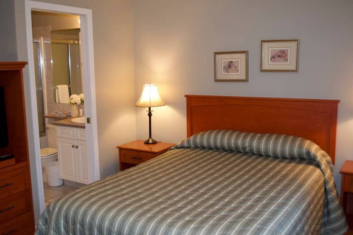 A hotel room with a bed, a nightstand with a lamp, a wooden headboard, a small table with a vase of flowers, and a bathroom visible through an open door.