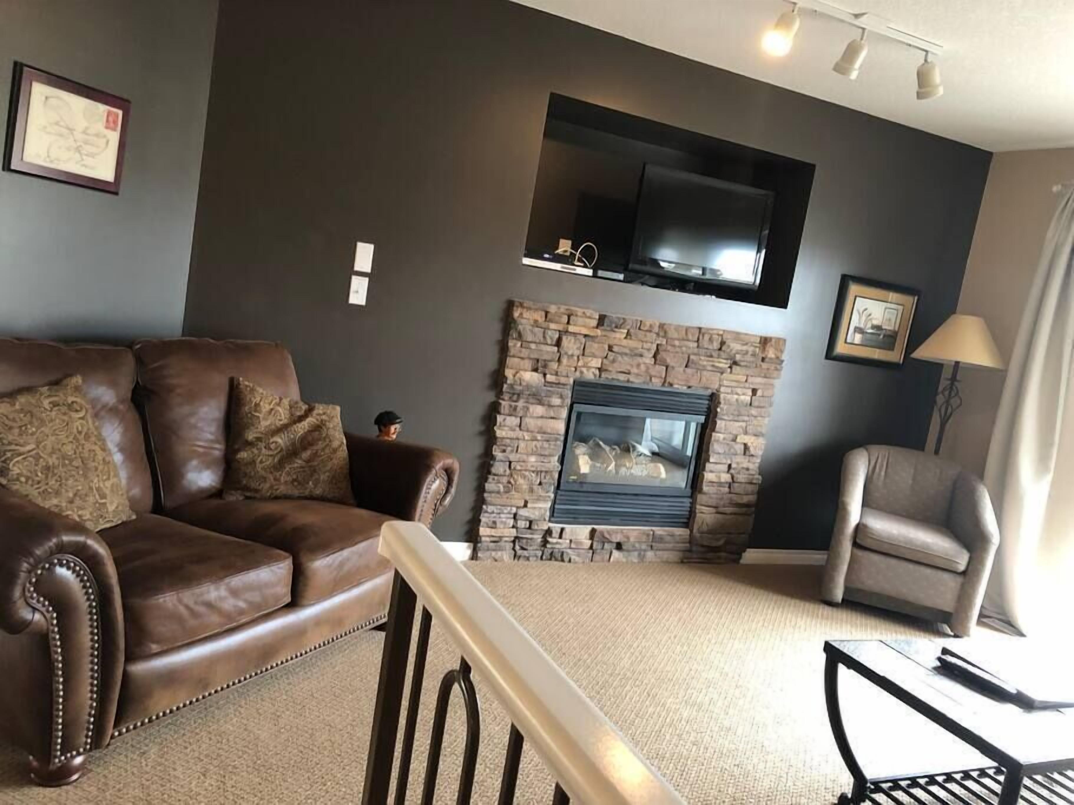 A living room with a brown leather sofa, a stone fireplace, a television mounted above it, and a grey armchair.