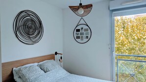 A bedroom with a bed, a wall-mounted circular decoration, a wall-mounted light fixture, and a window with a view of trees. - F2 garage,terrace,wifi,near town hall/train station/richter (Montpellier)