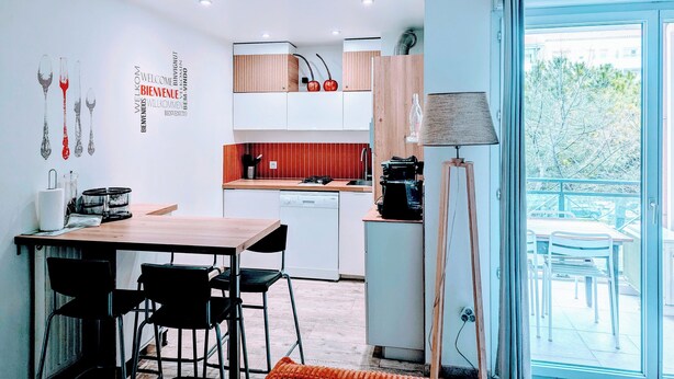 A modern kitchen with wooden cabinets, a dining area with a table and chairs, and a view of the outdoors through a glass door. - F2 garage,terrace,wifi,near town hall/train station/richter (Montpellier)