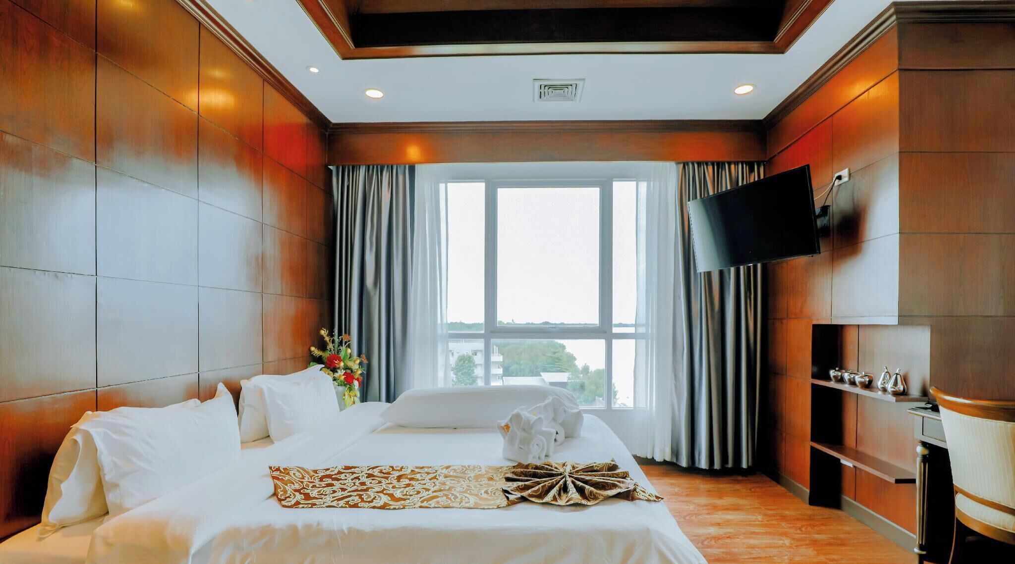 A hotel room with a large bed, a flat-screen TV, a window with curtains, and a vase of flowers.