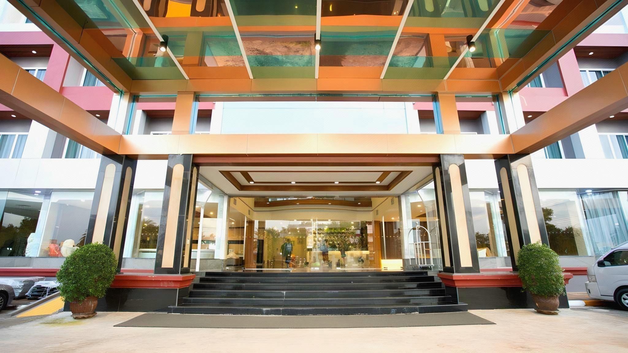 A hotel entrance with a glass facade, a staircase leading up to the entrance, and a parking area with cars.