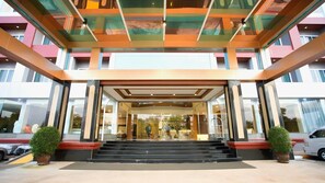 A hotel entrance with a glass facade, a staircase leading up to the entrance, and a parking area with cars. - CBD 2 Hotel (Surat Thani)