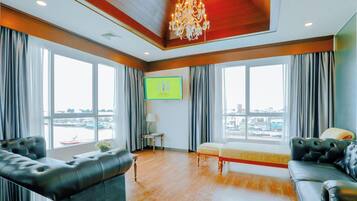 A hotel room with a large window, a chandelier, a sofa, a bench, and a TV screen.
