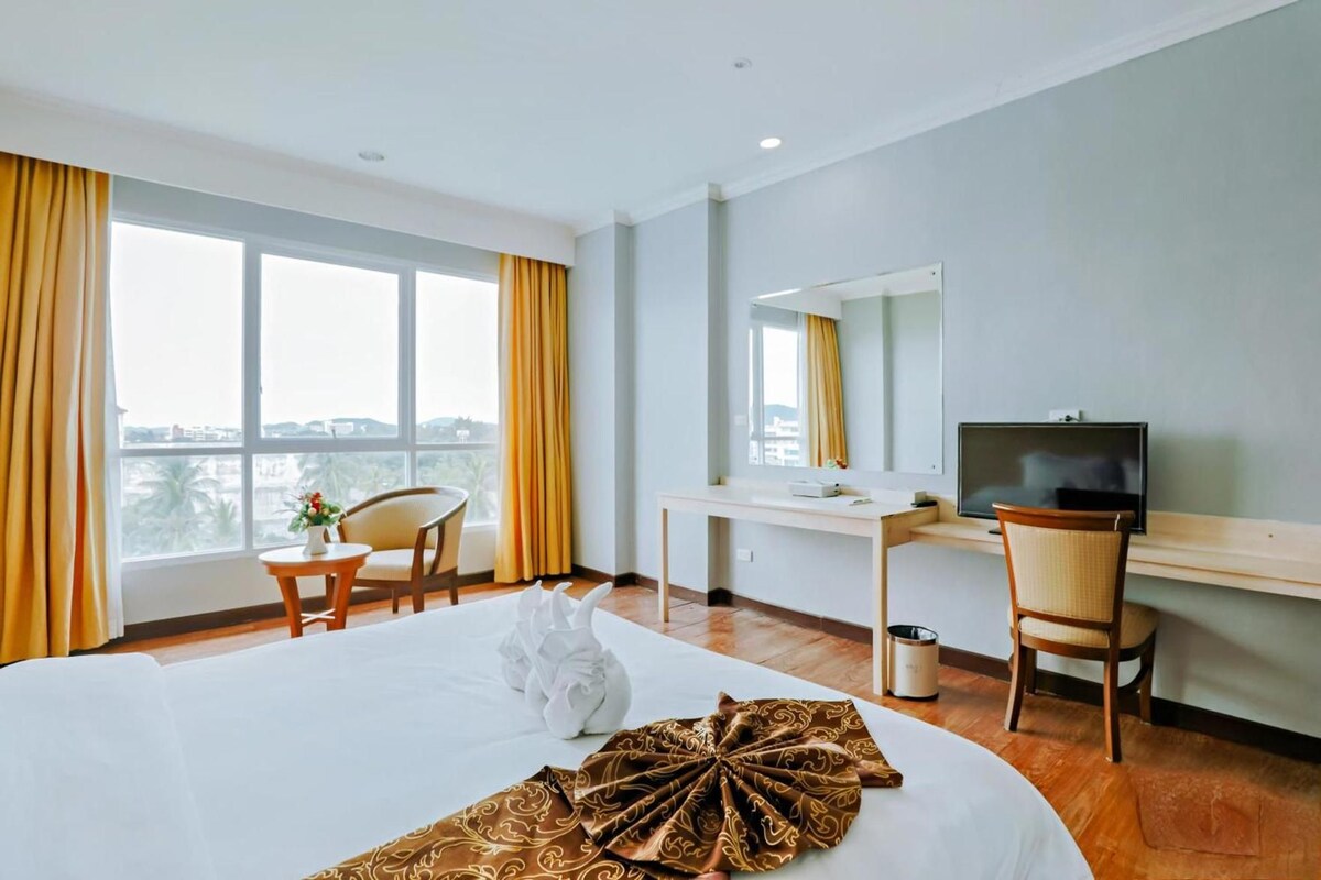 A hotel room with a bed, a desk, a chair, a television, and a view of the city through large windows.
