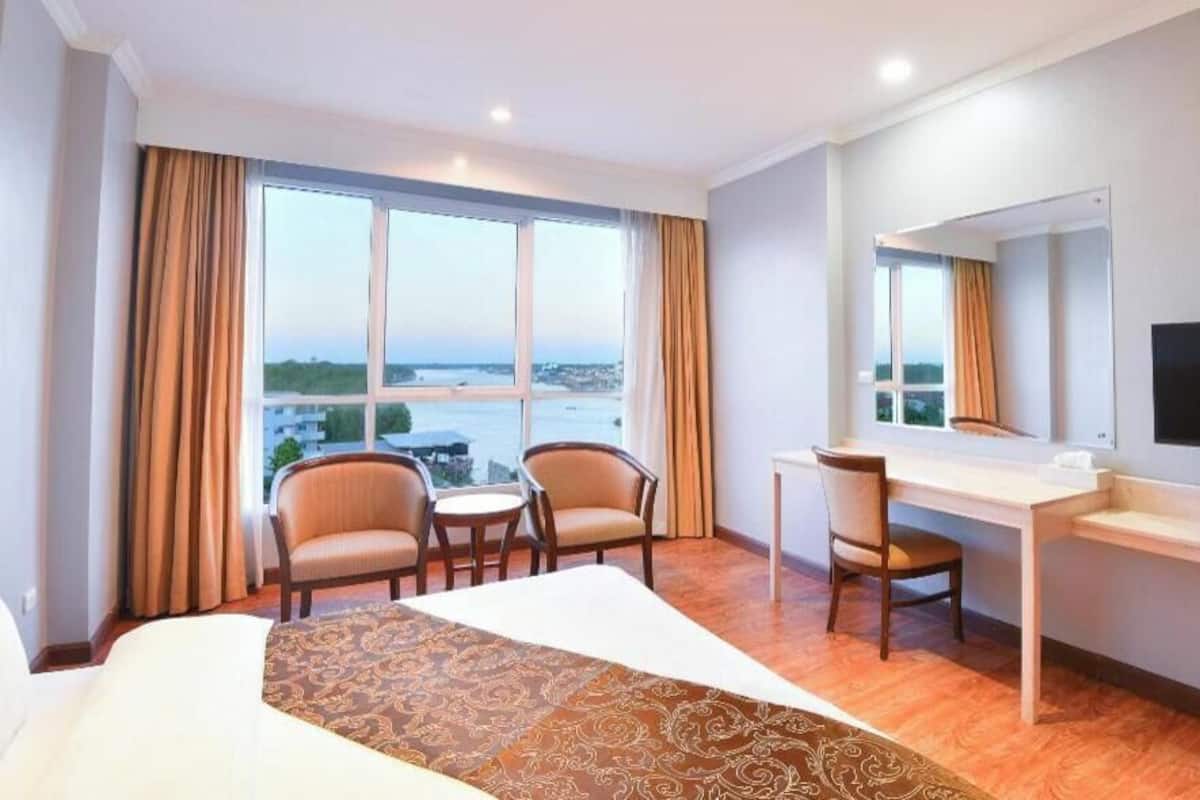 A hotel room with a bed, a desk, two chairs, a television, and large windows with curtains.