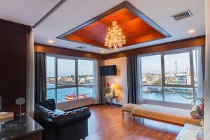 A hotel room with a large window, a chandelier, a sofa, a bench, and a flat-screen TV. - CBD 2 Hotel (Surat Thani)