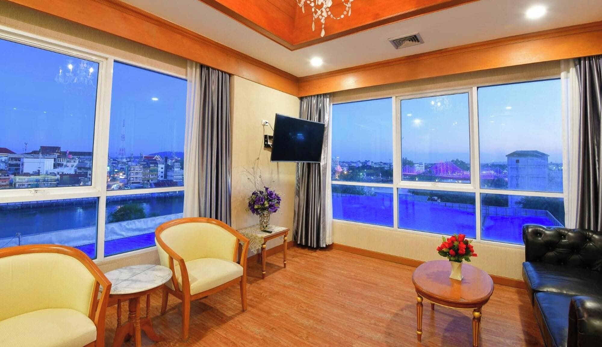 A hotel room with large windows, a flat-screen TV, a sofa, two armchairs, and a small table with a vase of flowers.