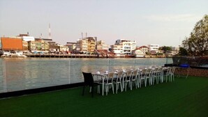 A waterfront dining area with chairs and tables, a grassy foreground, and buildings in the background. - CBD 2 Hotel (Surat Thani)