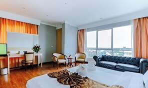 A hotel room with a bed, a sofa, a desk with a computer, two chairs, a small table with a vase of flowers, and a large window with curtains. - CBD 2 Hotel (Surat Thani)
