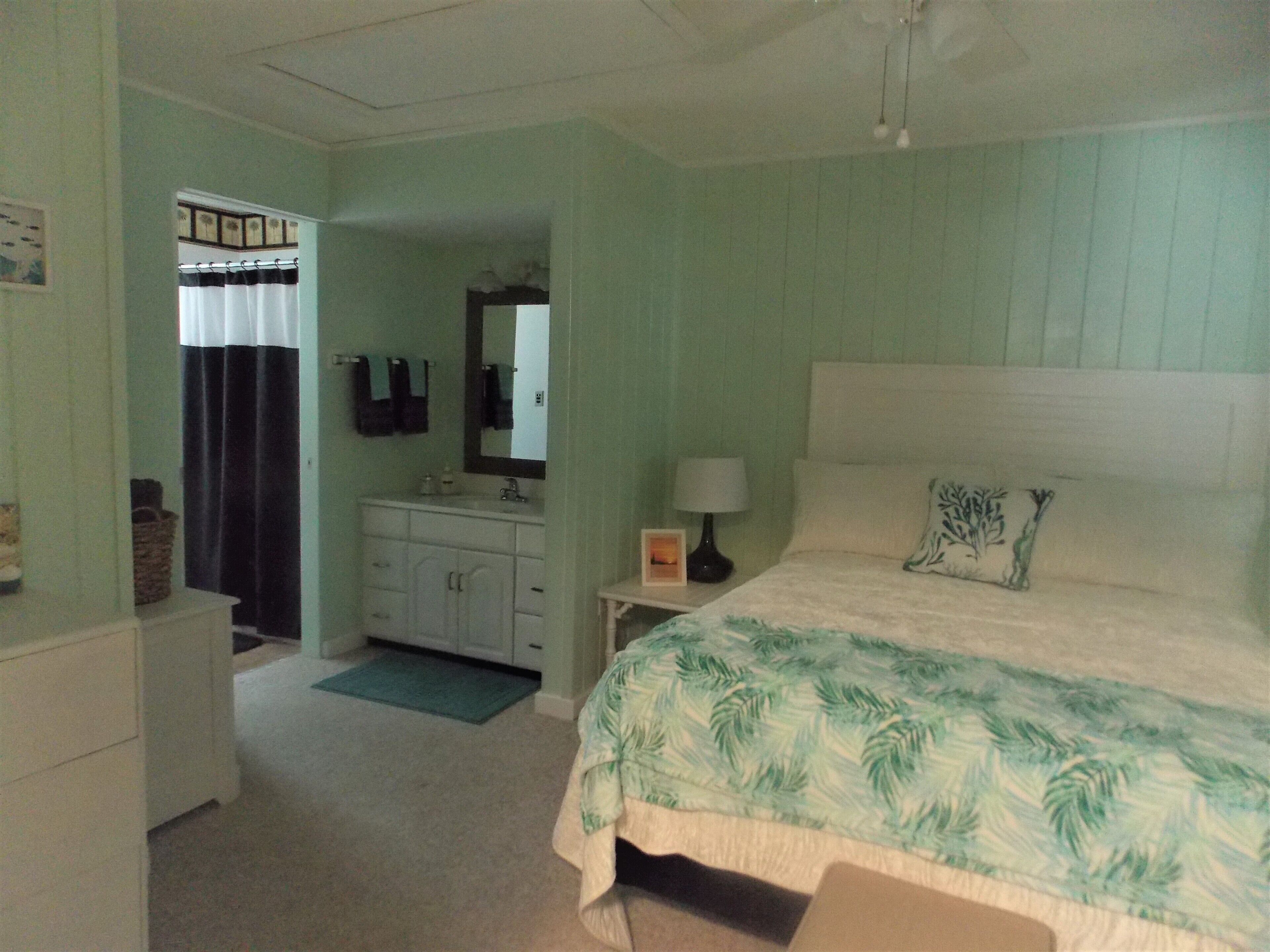 A bedroom with a bed, a nightstand, a dresser, a mirror, and a bathroom visible through an open door.