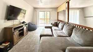 A living room with a flat-screen TV, wooden entertainment unit, grey sofa, and a sliding glass door leading to a balcony.
