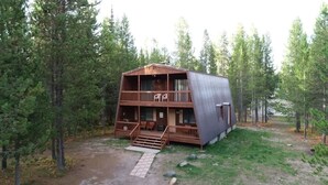 A two-story cabin with a wooden deck and a covered porch, surrounded by a forest.