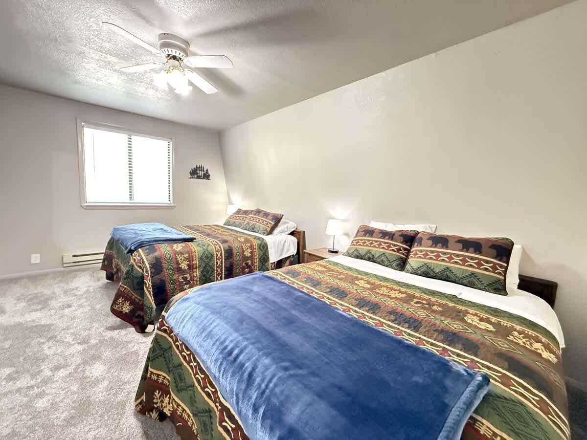 A bedroom with two beds, a ceiling fan, a window, and a wall-mounted heater.