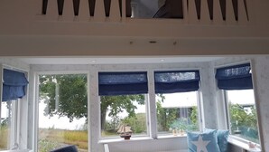 Quarto com teto alto, varanda com balaústres brancos e grandes janelas com cortinas azuis.