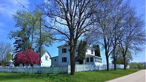 A white two-story house with a white picket fence, surrounded by green lawns and trees. -  Bloomin Pickets Farmhouse Stay OR Ladies Craft Retreat! (Monroe)