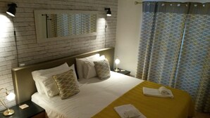 Hotellihuone, jossa on tiiliseinä, valkoisilla lakanoiden peitetty sänky, sängynpääty, yöpöytä lampun kanssa ja kuviollinen verho.