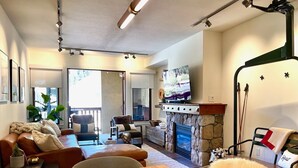 A living room with a stone fireplace, a television on a stand, and a dining area with a wooden table. - The Village at Palisades Tahoe Bldg 1 - Chic Condo with Prime View (Olympic Valley)