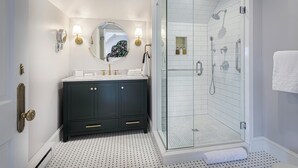 A modern bathroom with a dark vanity, a round mirror, a glass-enclosed shower, and a tiled floor. - The West Lane Inn - King Room (Ridgefield)