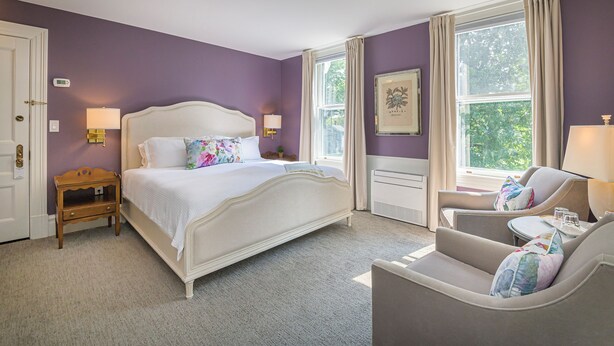 A bedroom with a large bed, a nightstand, a chair, and a window with curtains. - The West Lane Inn - King Room (Ridgefield)