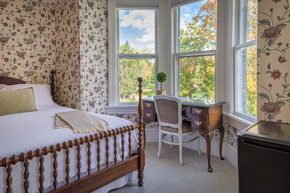 Une chambre à coucher avec un lit, un bureau et une vue sur les arbres, visible à travers de grandes fenêtres.