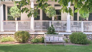 A covered porch with a stone wall, a bench, hanging planters, and a view of the house. - The West Lane Inn - Deluxe Fireplace King (Ridgefield)