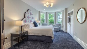 A bedroom with a large bed, two bedside tables, a dresser, and a mirror. There are windows providing natural light and a view of greenery outside. - The West Lane Inn - Deluxe Fireplace King (Ridgefield)