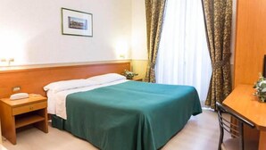 Una habitación de hotel con cama, escritorio, silla, mesita de noche y un cuadro en la pared.