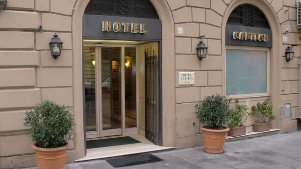 Entrada de un hotel con puerta de vidrio, un cartel que dice "HOTEL CAPITOL" y dos plantas en macetas en la acera.