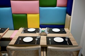 A dining area with booths and tables set for four, each with a place setting including a plate, fork, spoon, and glass. - Sun City (New Delhi)