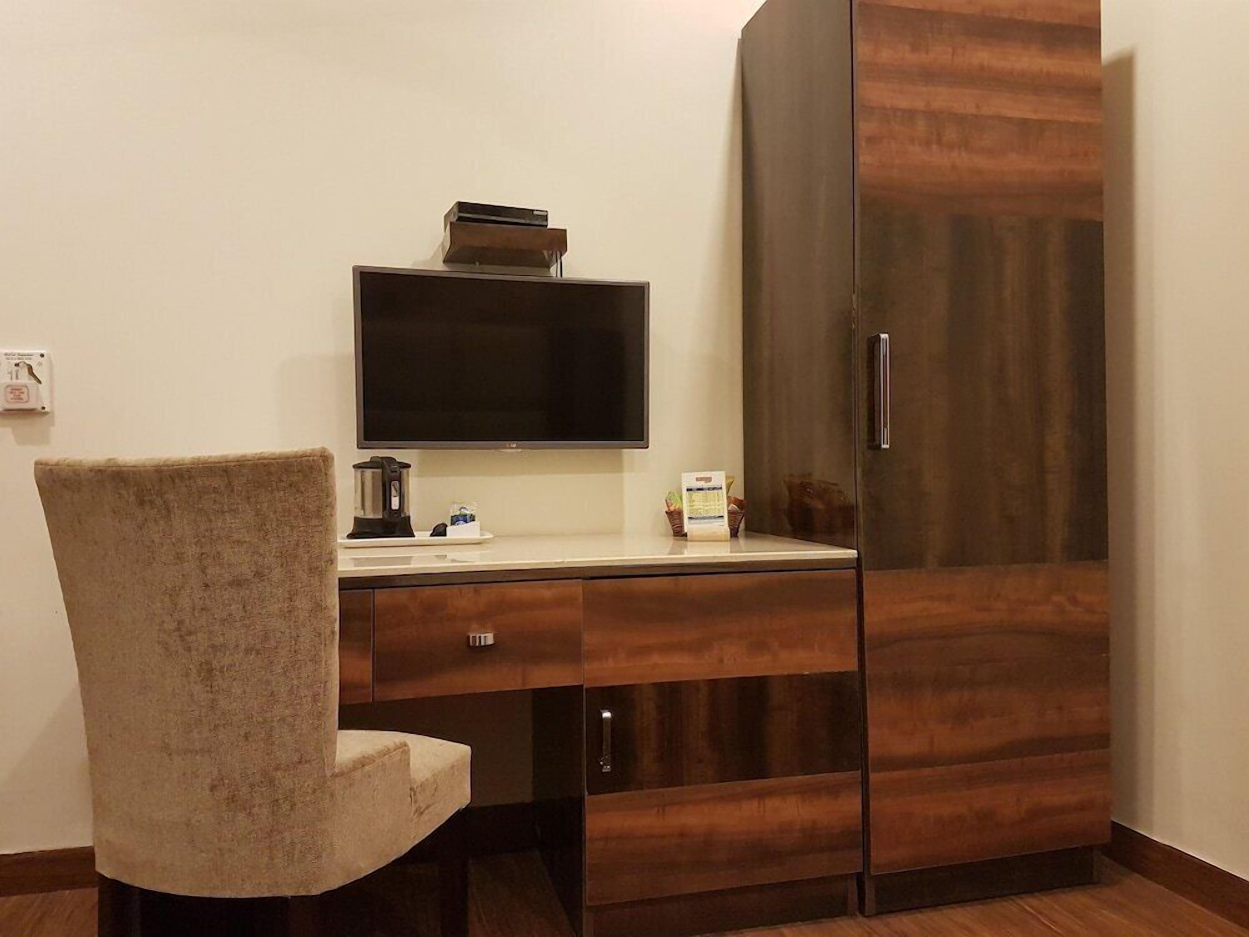 A room with a wooden wardrobe, a flat-screen TV, a coffee maker, and a chair.