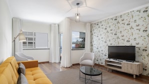 A modern living room with a sofa, a coffee table, a TV on a wooden stand, and a window with curtains. - By the sea, Reis Magos IV (Caniço)