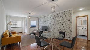 A modern living room with a sofa, a glass dining table with chairs, a TV, and a view of the sea through a window. - By the sea, Reis Magos IV (Caniço)