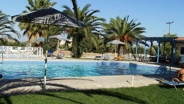 A swimming pool with lounge chairs and umbrellas, surrounded by palm trees and a gazebo. - Zeus (Samos)