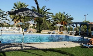 A swimming pool with lounge chairs and umbrellas, surrounded by palm trees and a gazebo. - Zeus (Samos)