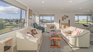 A spacious living room with large windows, a green chair, a beige sofa, and a wooden coffee table. - Pauanui Hangar Retreat - Pauanui Holiday Home (Pauanui)