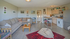 Un espacio habitable compacto con un sofá, una mesa de centro y una alfombra roja. Se ve una lavadora y una zona de cocina al fondo.