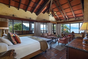 A spacious room with a large bed, a seating area with a table, and large windows offering a view of the outdoors. - Atitlan (Sololá)