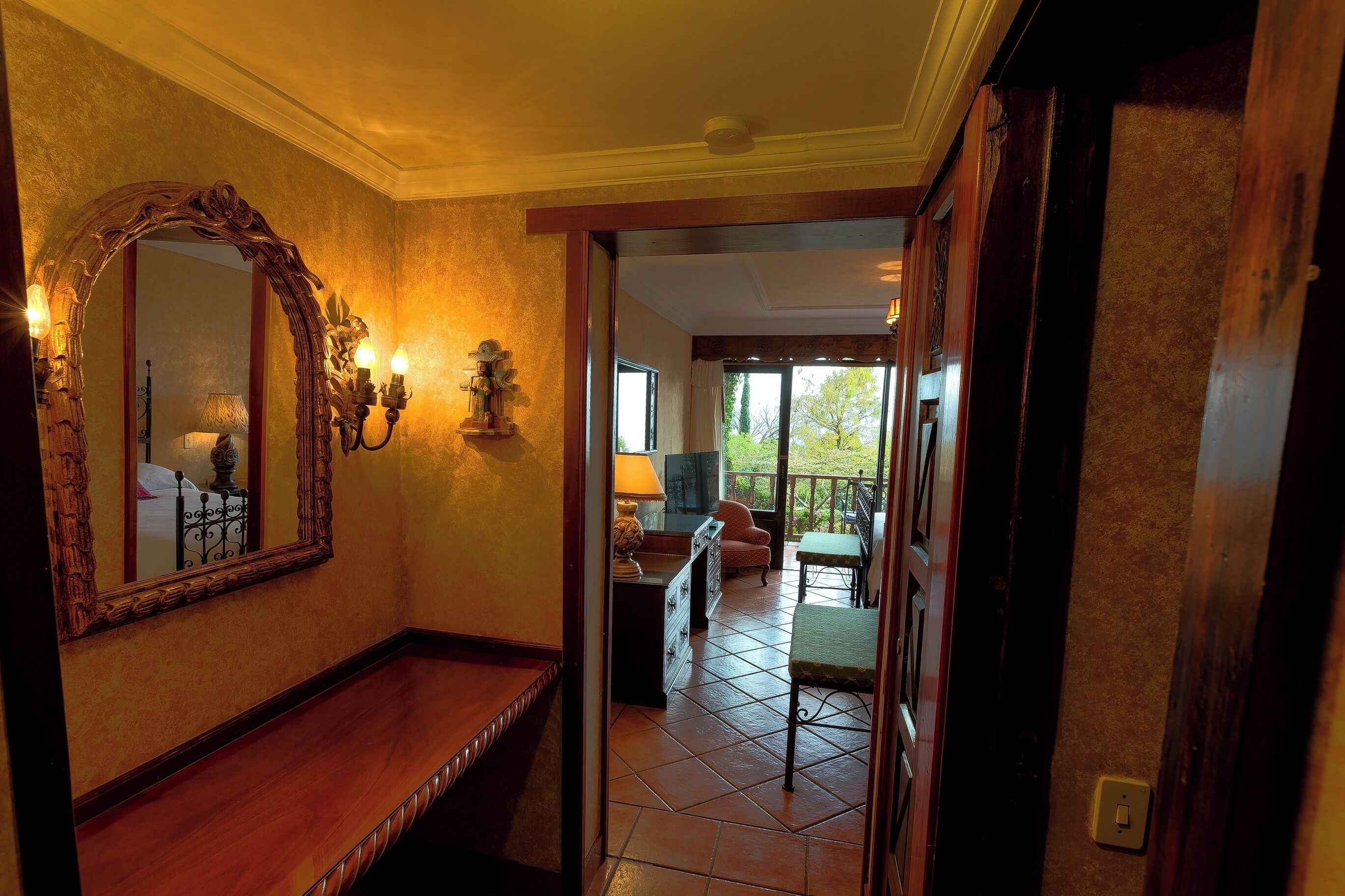 Un pasillo con un banco de madera, un espejo grande y ornamentado, apliques en la pared y vistas a un balcón con mobiliario exterior.