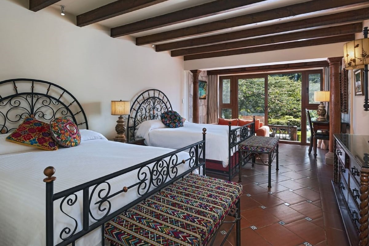 A room with two beds, a small table, and a view of greenery outside.