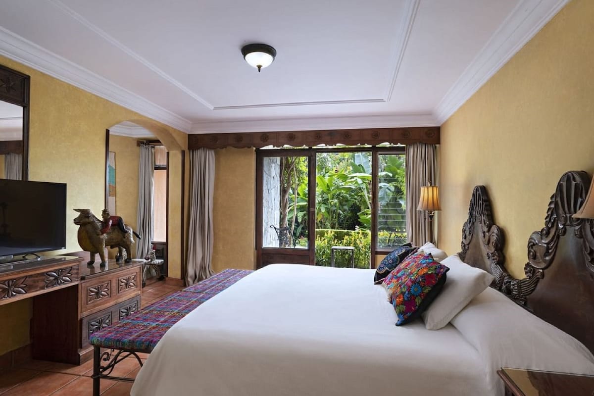 A hotel room with a large bed, a television, a desk with drawers, and a view of greenery outside.