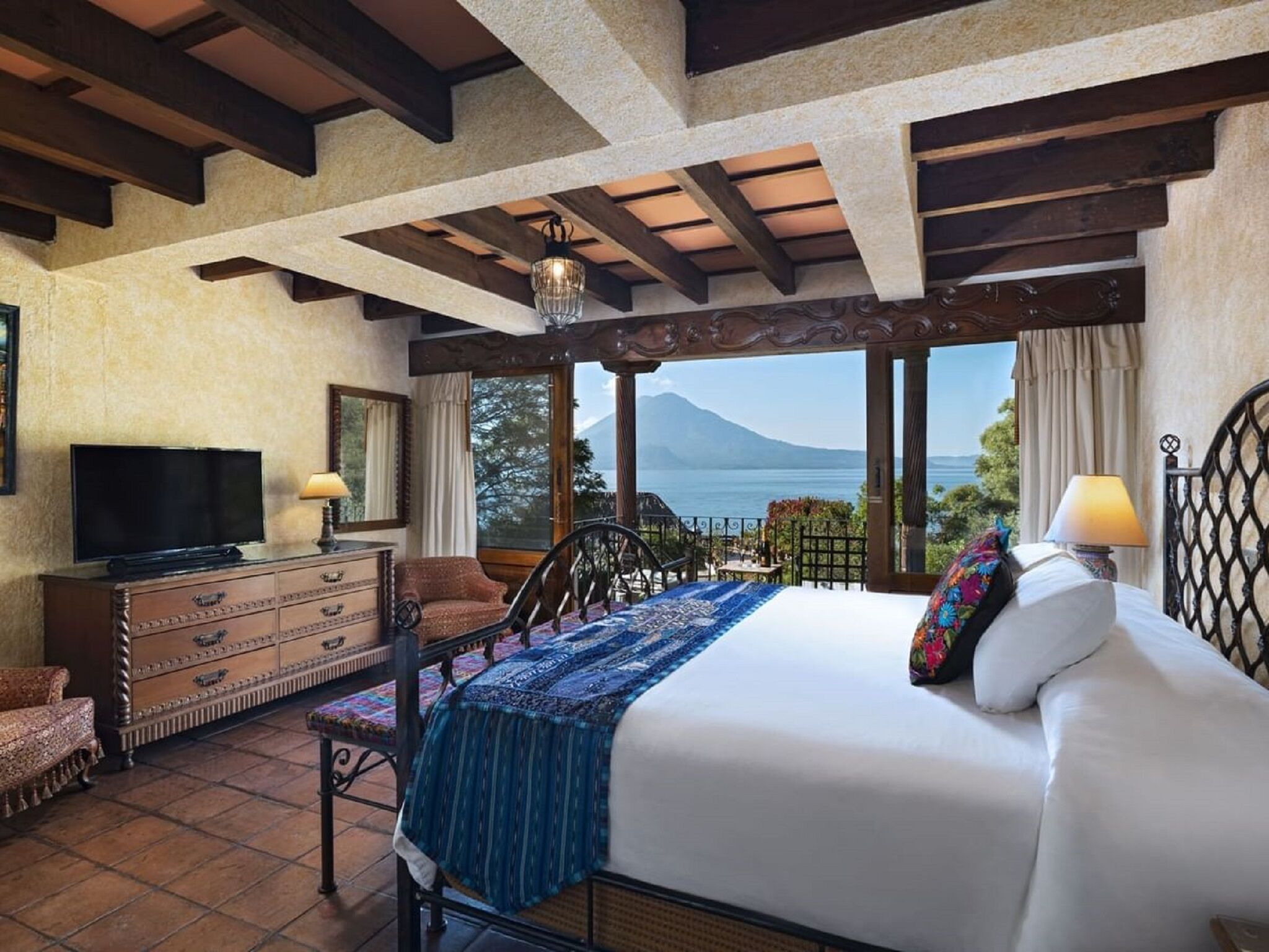 Un dormitorio con cama, televisor, cómoda, silla y balcón con vistas a la montaña y al mar.