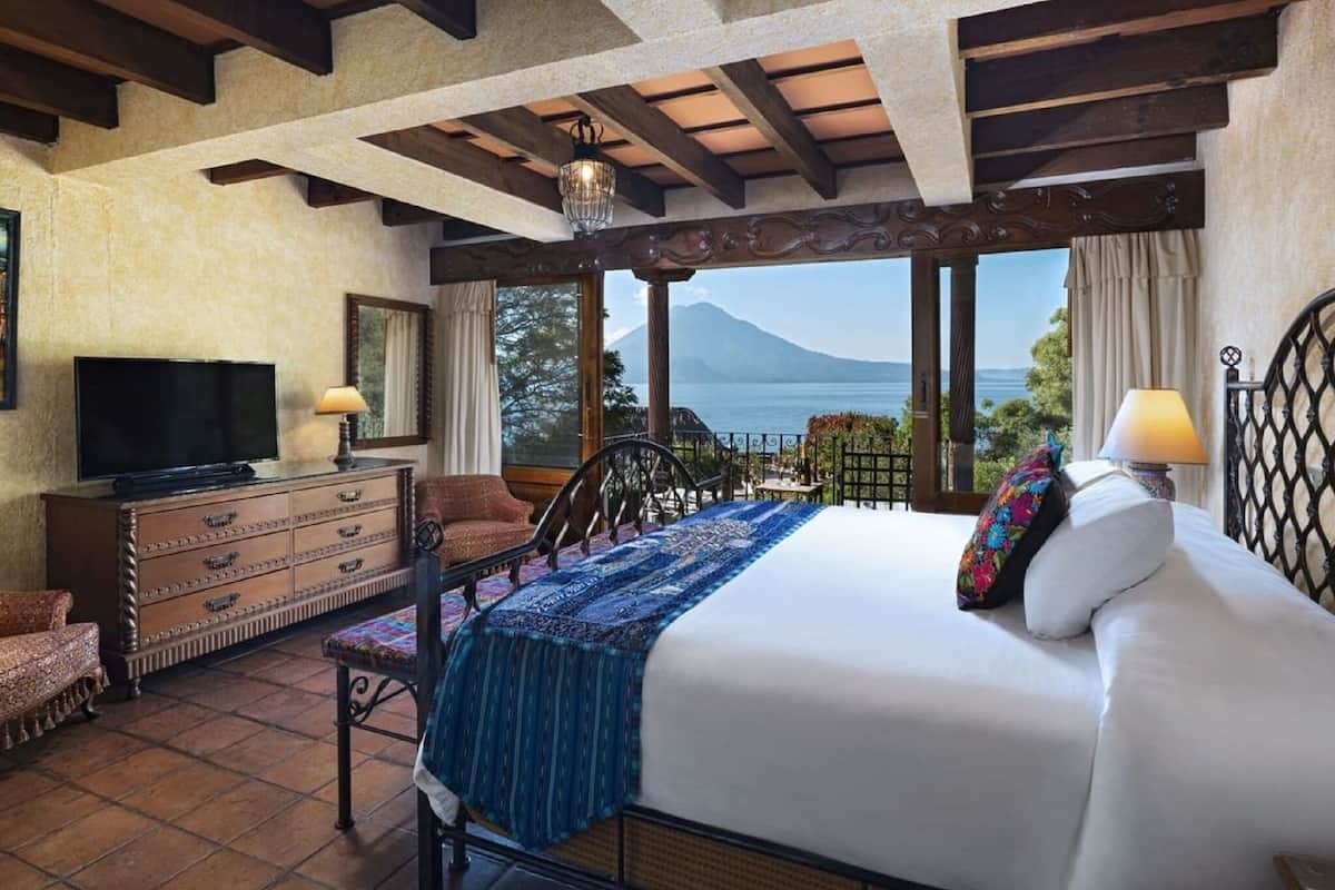 A bedroom with a bed, a television, a dresser, a chair, and a balcony with a view of a mountain and sea.