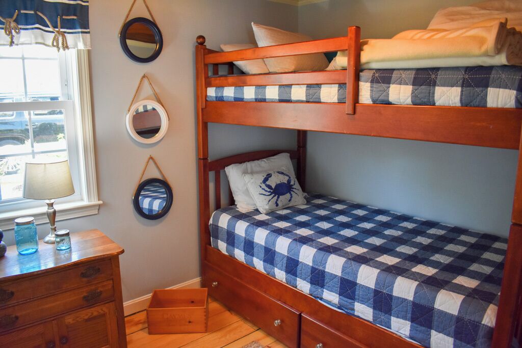 Een stapelbed met een geruit sprei, een houten dressoir met lades en een raam met gordijnen.