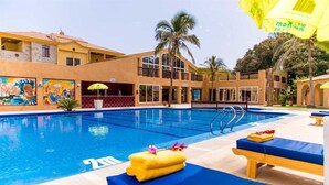 A swimming pool with lounge chairs and towels, surrounded by buildings with murals and palm trees. - Tropic Gardens Hotel (Bakau)