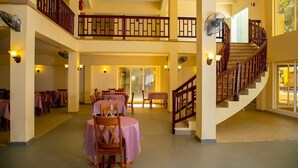 A spacious indoor area with multiple dining tables and chairs, a staircase, and a fan on the upper level. - Tropic Gardens Hotel (Bakau)