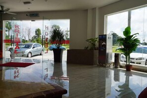 Zona de recepción de un hotel con puerta de vidrio que da al exterior, un mostrador de recepción y una planta en maceta.