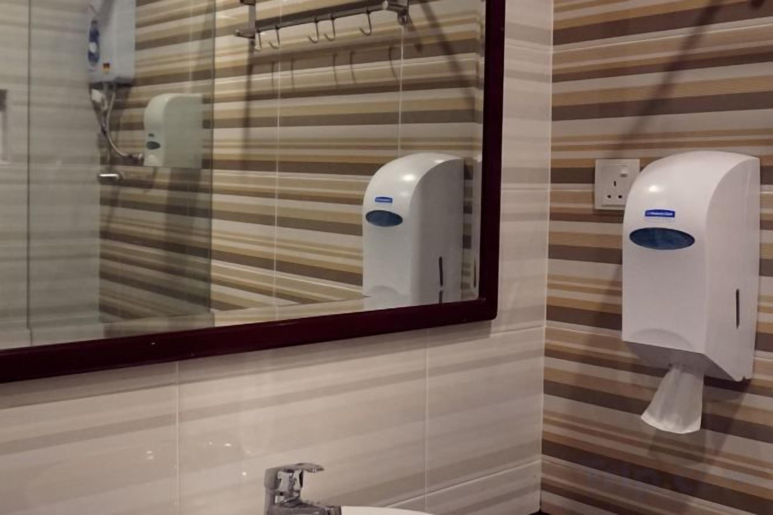 Une salle de bain avec un mur recouvert de carreaux à rayures, un miroir, un lavabo et un distributeur de gel hydroalcoolique.